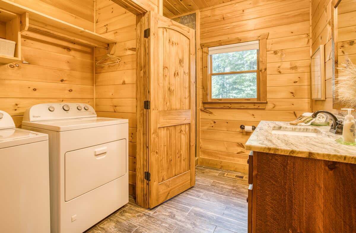 A wooden cabin interior with a washing machine, a wooden door, a window, and a countertop with toiletries.