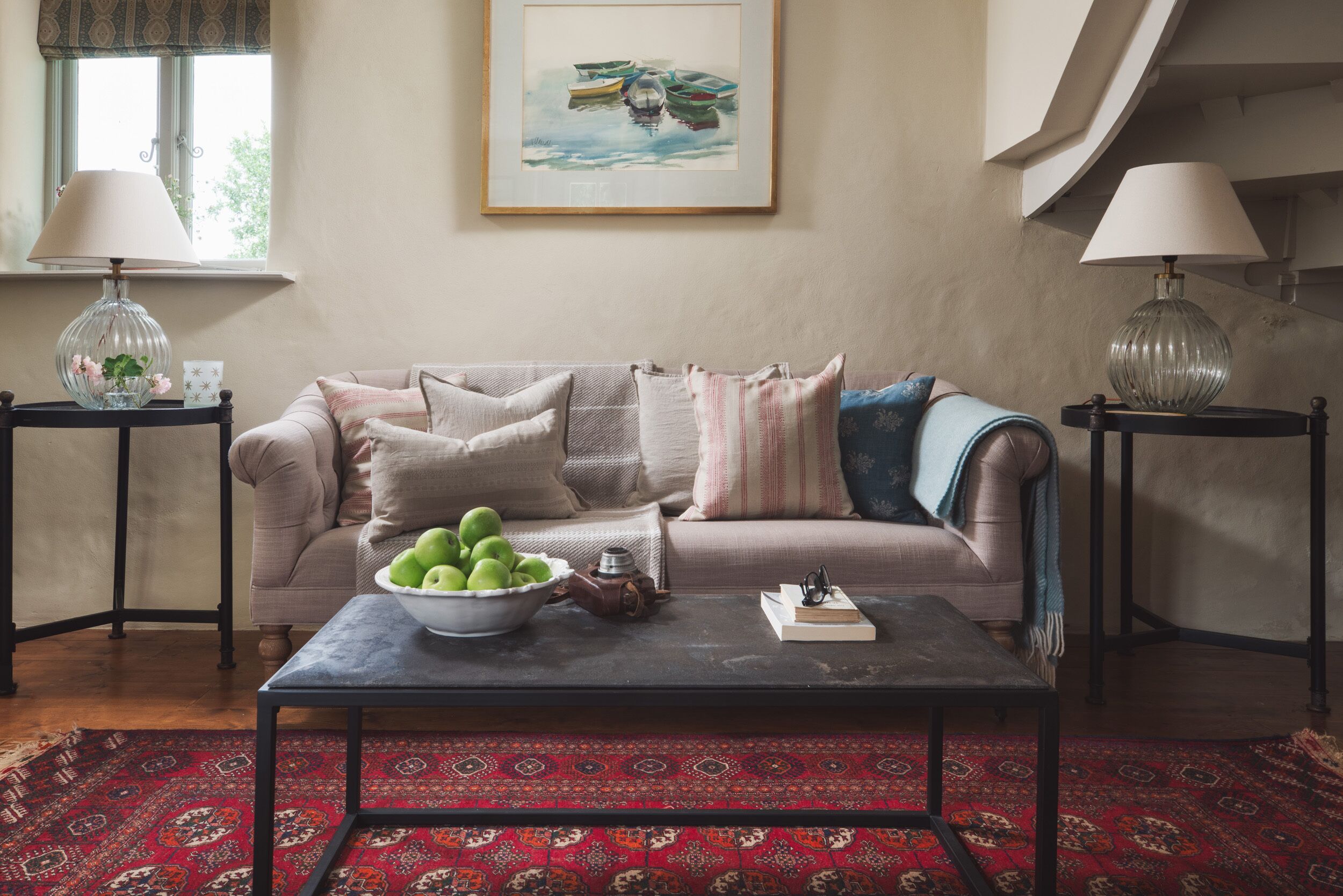 A living room with a sofa, two side tables, a coffee table with a bowl of apples, and a framed painting on the wall.