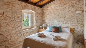 Une chambre avec des murs en pierre, un plafond en bois, un lit recouvert d’une courtepointe à rayures, une table de chevet avec une lampe et une fenêtre donnant sur un paysage verdoyant.