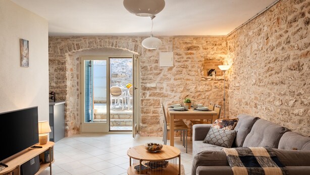 Un salon chaleureux avec un mur en pierre, un canapé, une table à manger avec des chaises et un téléviseur posé sur un meuble.