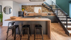 A modern kitchen with a central island, three bar stools, a stone accent wall, and a staircase with a metal railing. - La Longère du Verger, Maison de 200m2 Avec Piscine Intèrieure, et Plage à Pied (Trégunc)