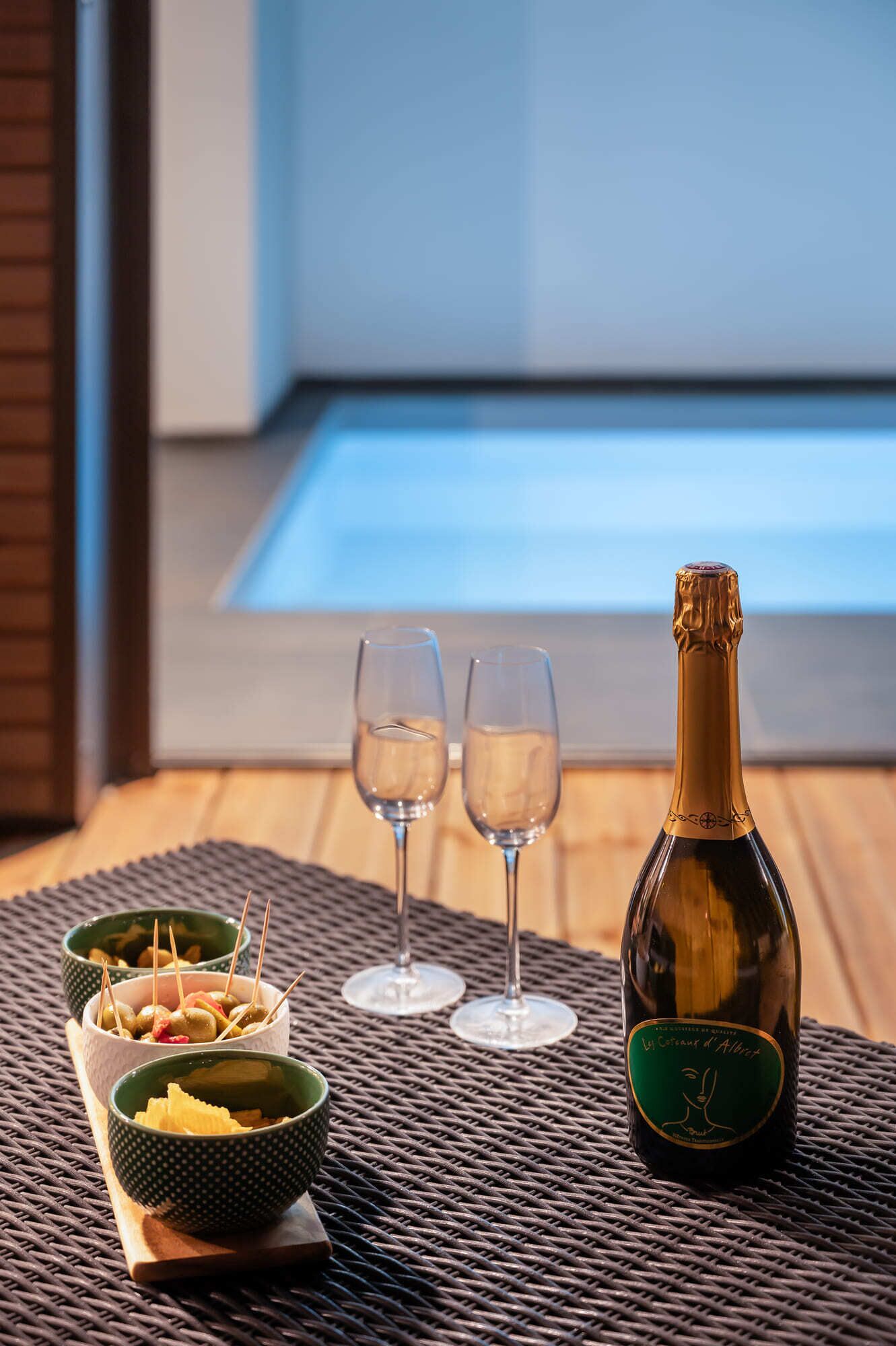 Une bouteille de champagne, deux verres et des bols remplis d’olives et de crackers, le tout posé sur une table d’où l’on peut admirer une piscine.