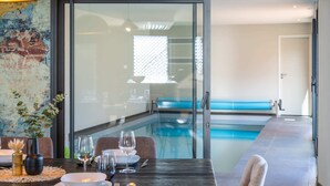 A dining area with a table set for two, a glass partition, and a view of a swimming pool. - La Longère du Verger, Maison de 200m2 Avec Piscine Intèrieure, et Plage à Pied (Trégunc)