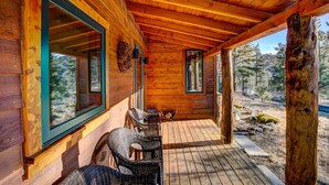 A wooden cabin with a covered porch, wicker furniture, and a view of the surrounding forest. - Evergreen View - A Modern Mountain Retreat (Estes Park)