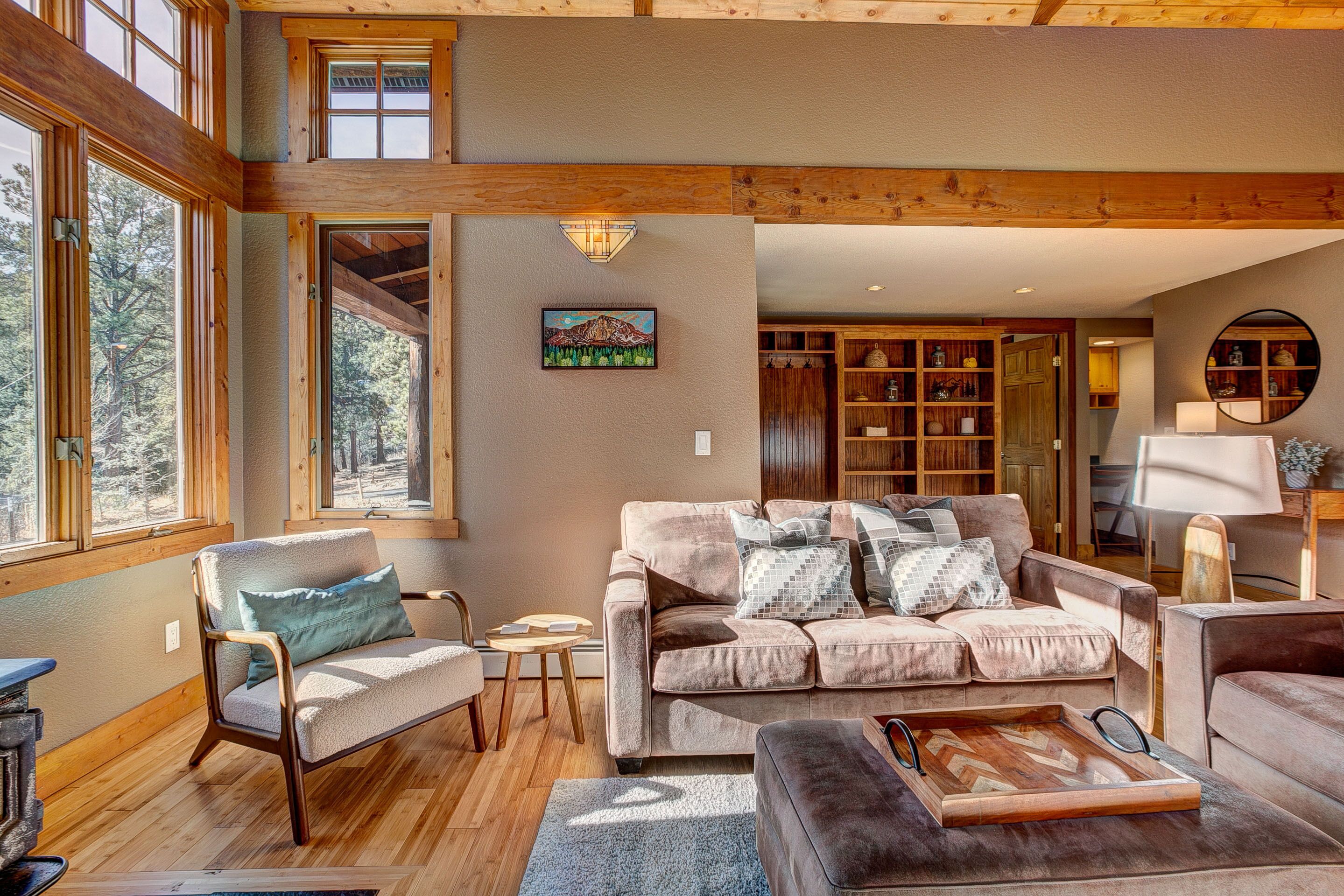 A cozy living room with a large window, wooden beams, a sofa, a chair, and a fireplace.