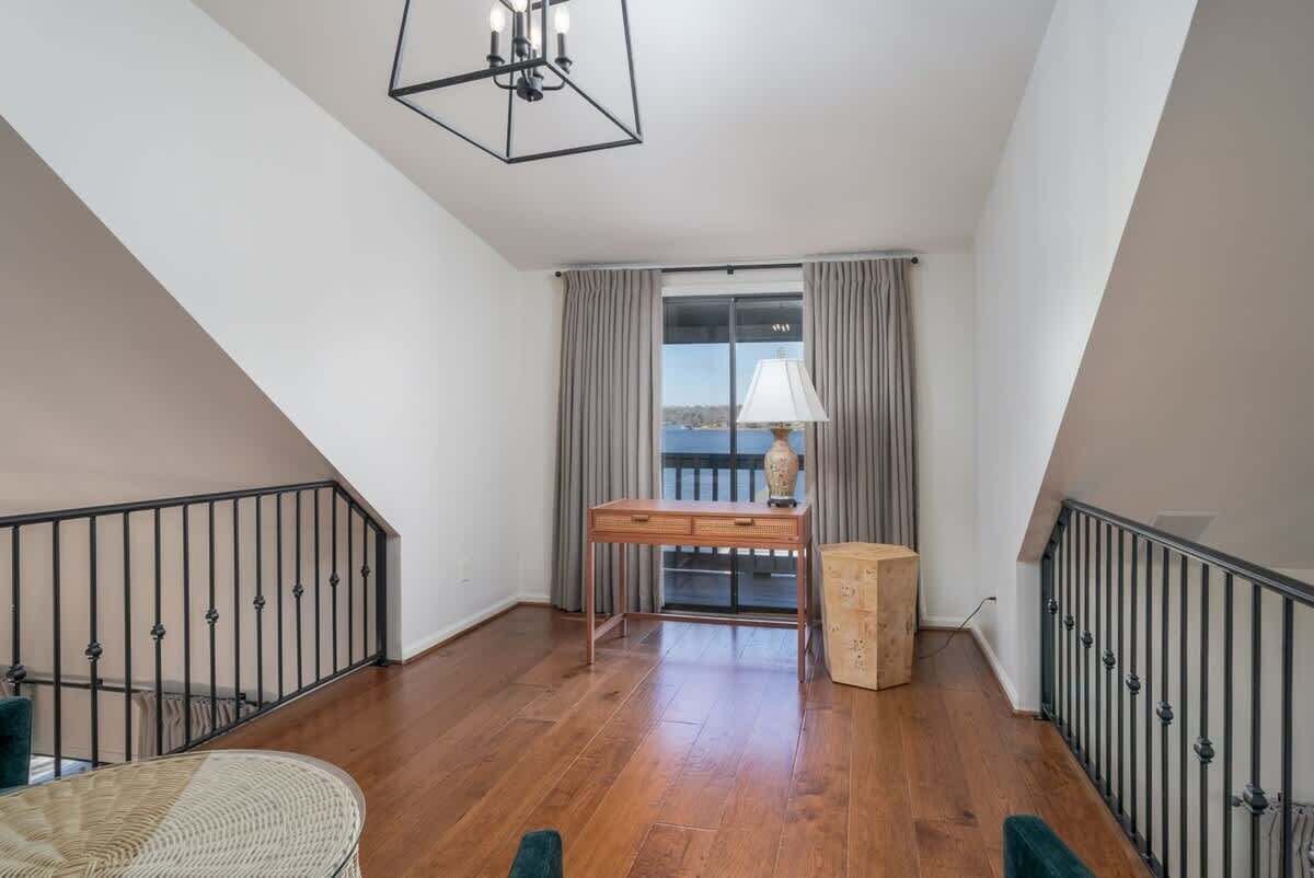 A spacious room with a wooden floor, a desk with a lamp, and a view through a large window.