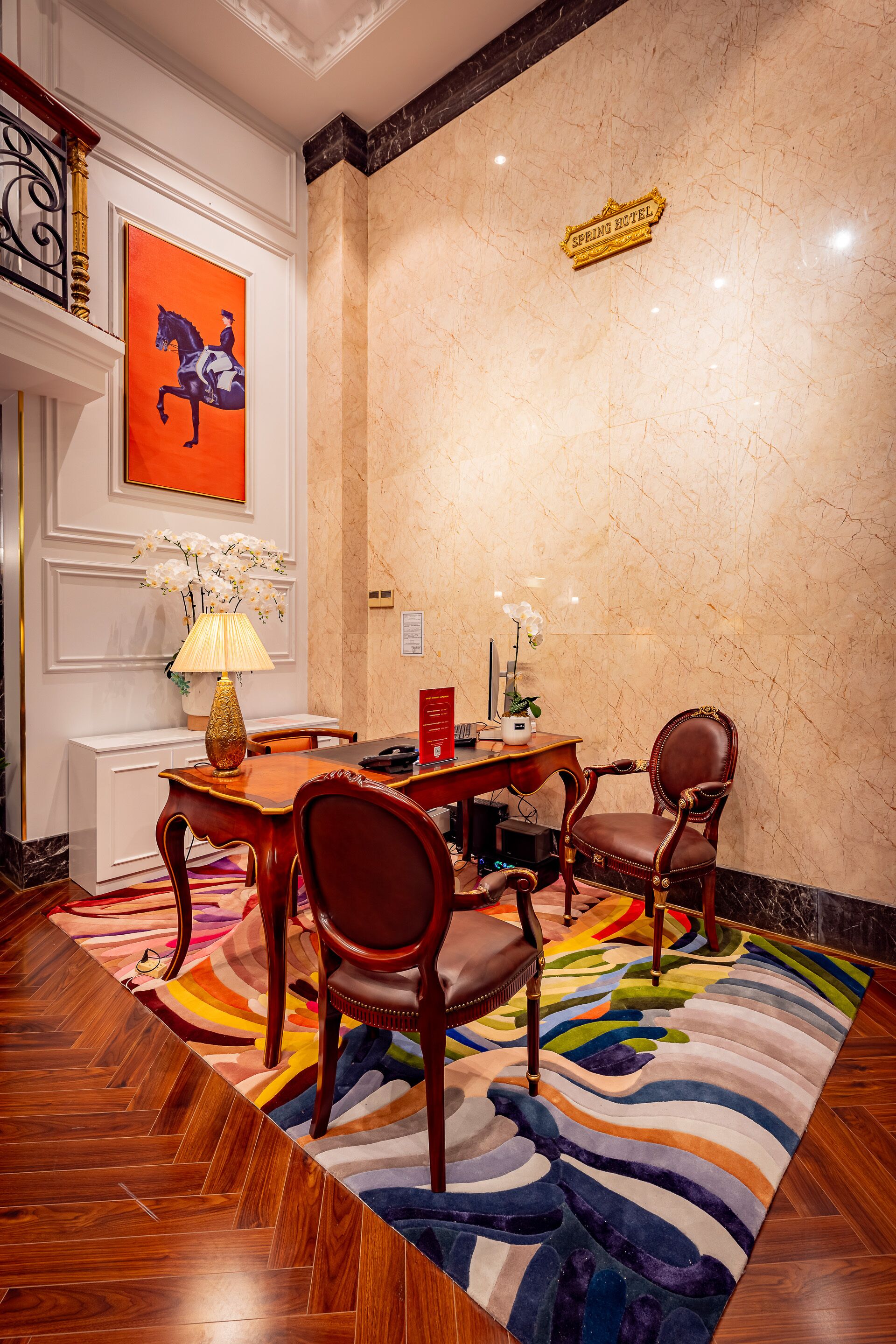 A hotel lobby with a marble wall, a red and white painting, a sign reading "SPRING HOTEL", a wooden reception desk, a chair, a table, and a colorful rug.