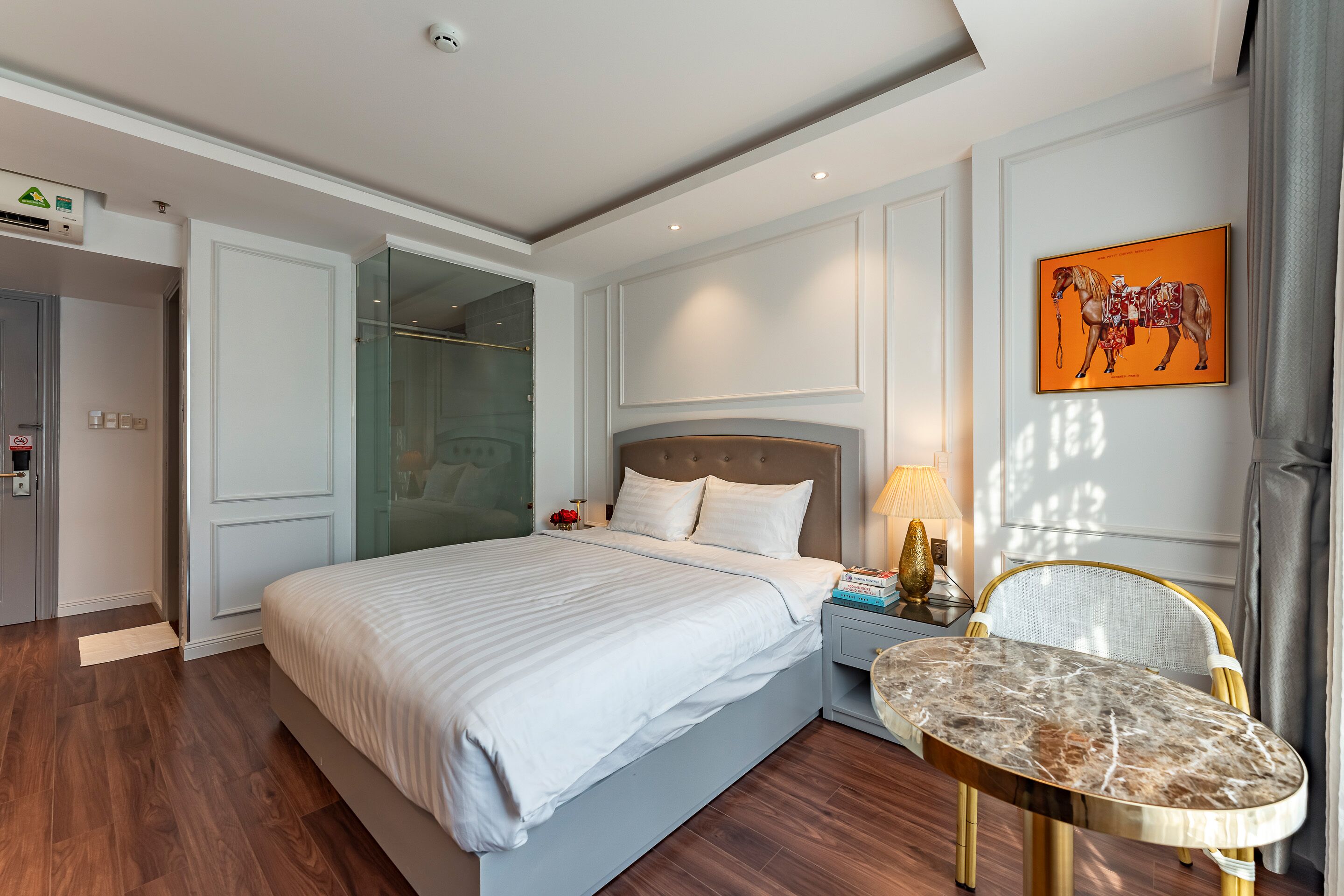 A modern hotel room with a large bed, a marble side table, a glass shower enclosure, and a framed artwork on the wall.