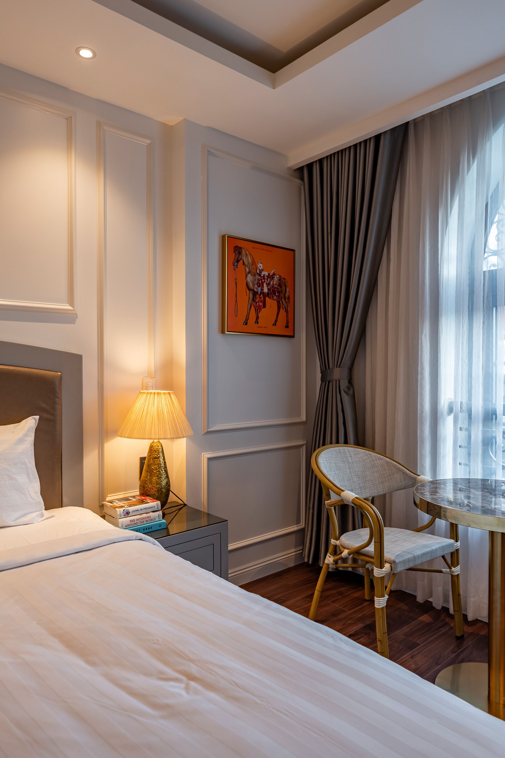 A hotel room with a bed, a bedside table with a lamp, a chair, a painting on the wall, and a window with curtains.