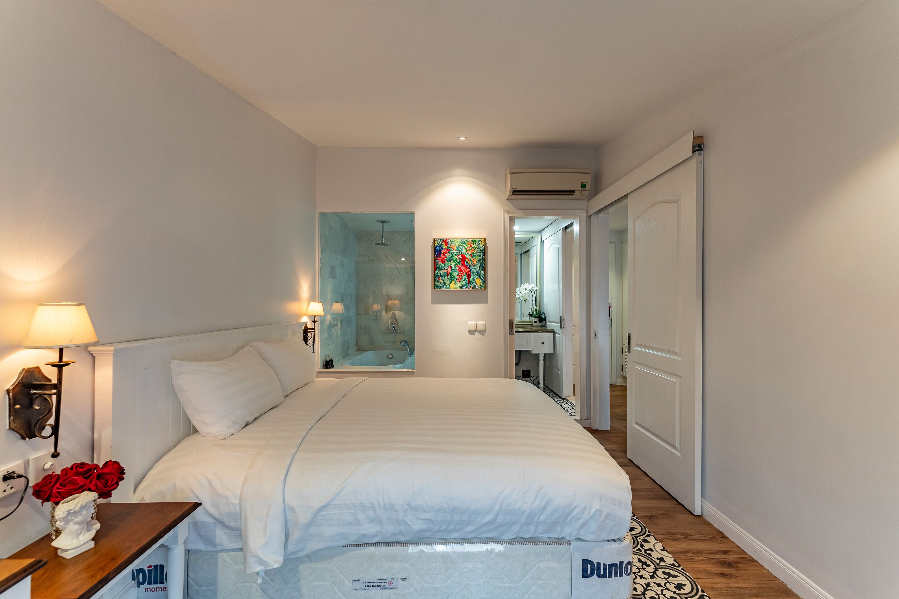 Ein modernes Hotelzimmer mit einem großen Bett, einem Nachttisch mit Lampe, einem kleinen Tisch mit roten Rosen und einem sichtbaren Duschbereich hinter einer Glastür.