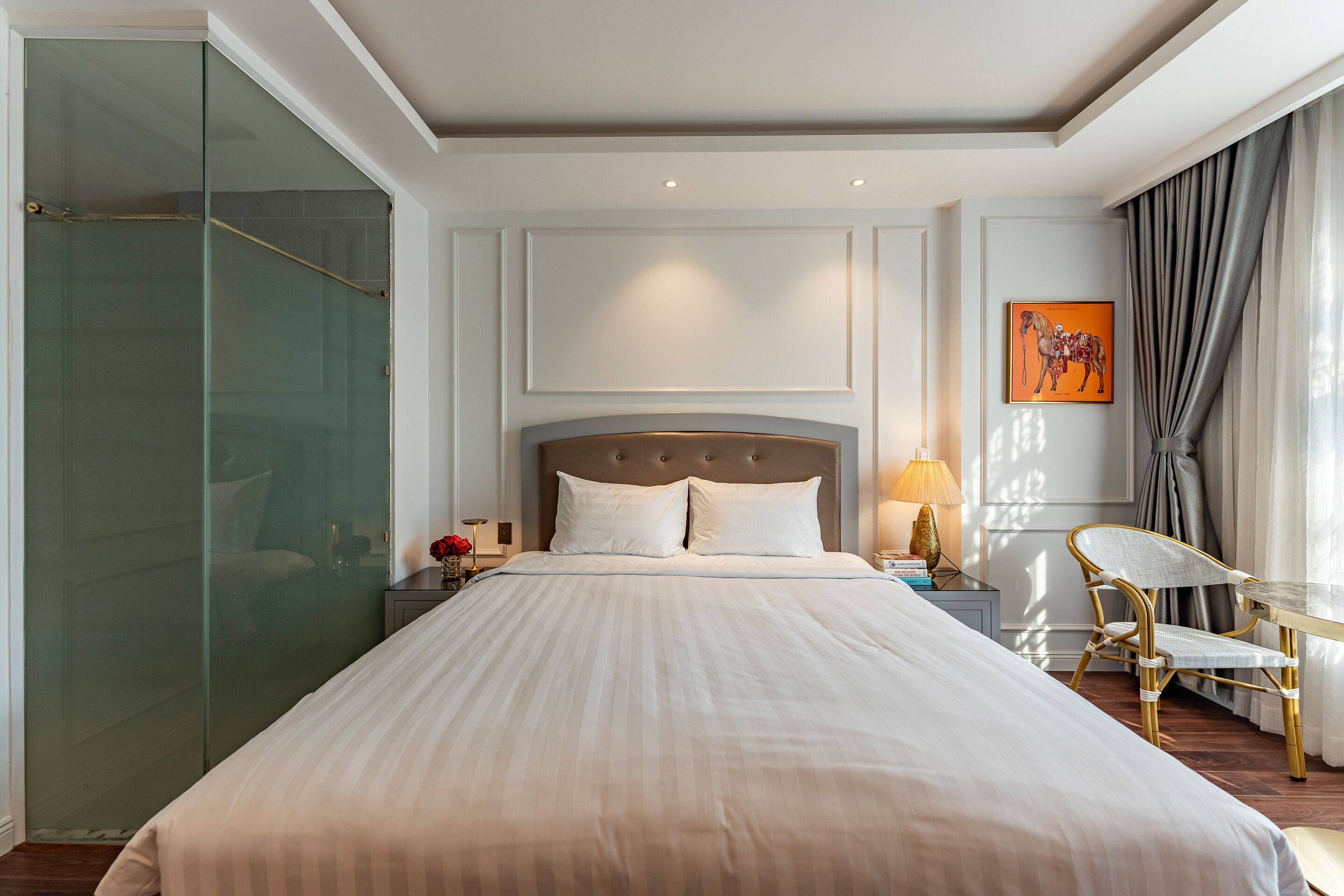 A hotel room with a large bed, a glass shower enclosure, a nightstand with a lamp, a chair, and a painting on the wall.