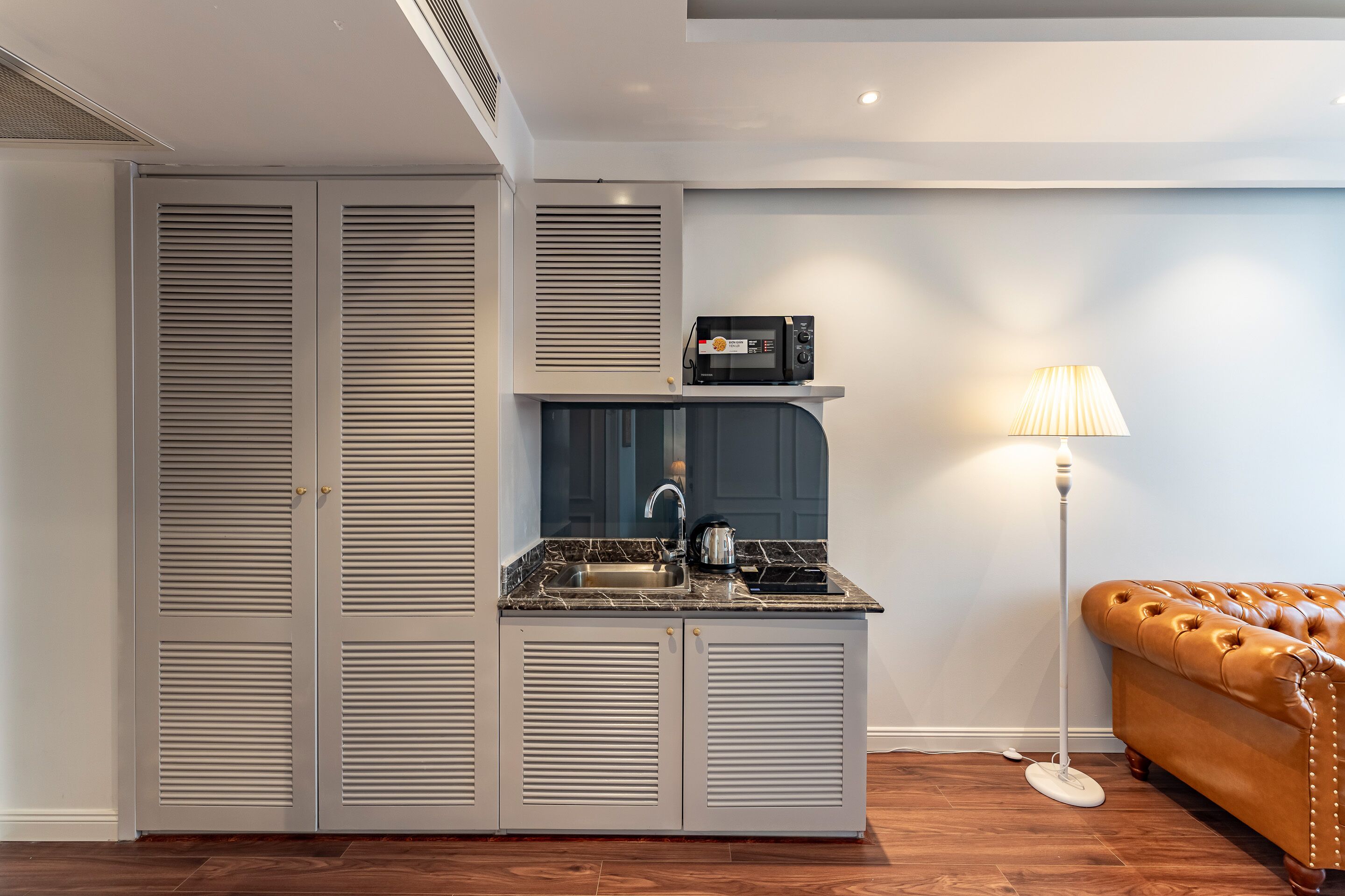 A compact kitchen with a built-in microwave, a kettle, and a sink. There is a brown leather sofa and a floor lamp in the adjacent living area.