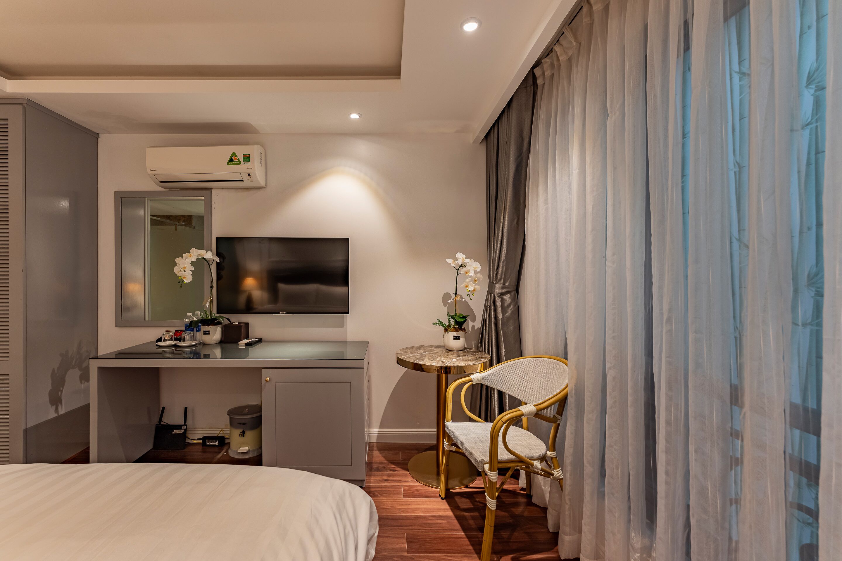 A modern hotel room with a bed, a desk with a flat-screen TV, a small table with a plant, and a chair.