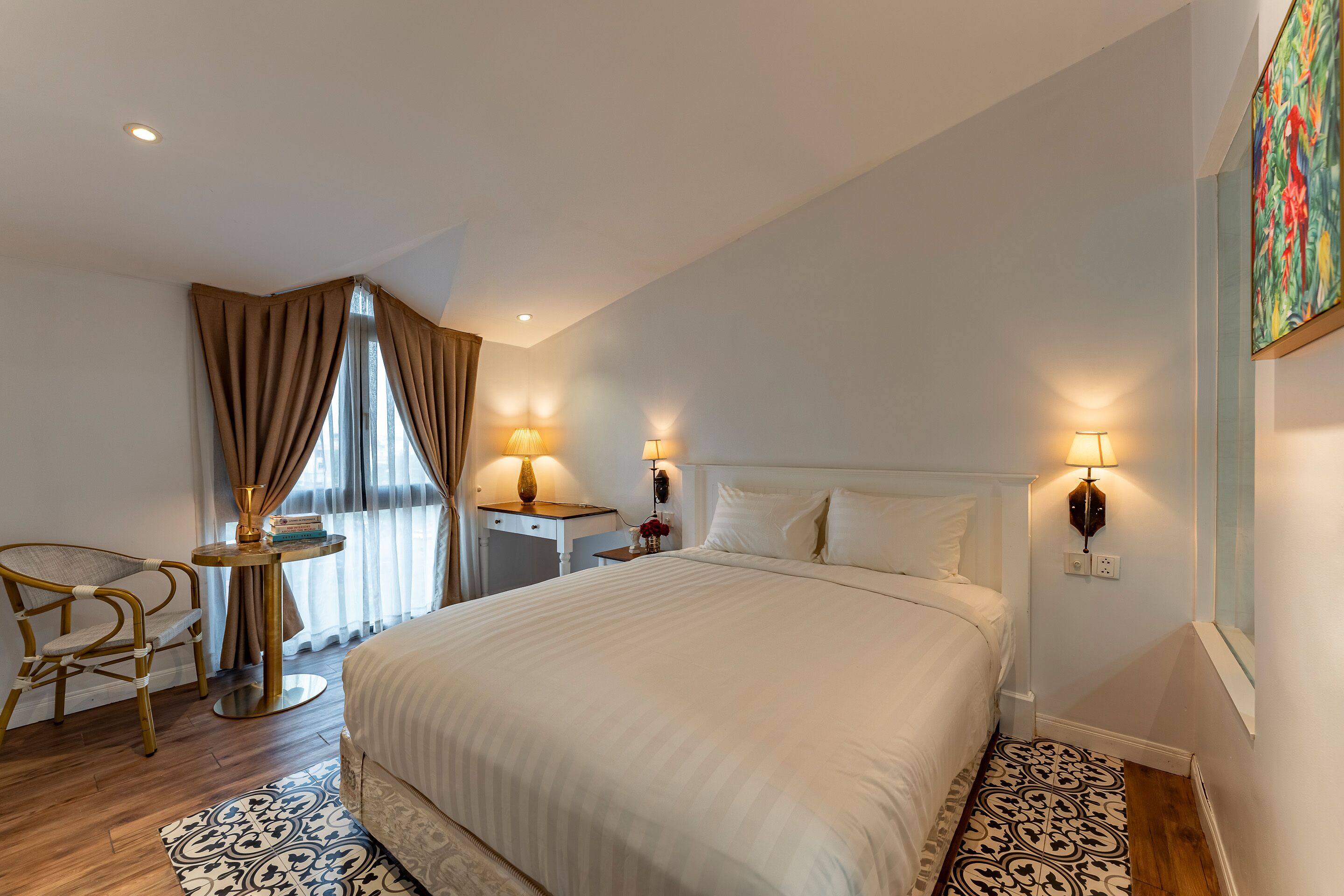 A hotel room with a large bed, two bedside tables with lamps, a chair, a window with curtains, and a painting on the wall.