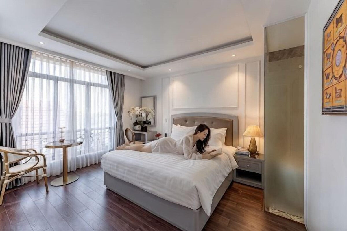 Habitación de hotel con una cama grande, una mesita de noche, una silla, una pequeña mesa y un ventanal con cortinas.