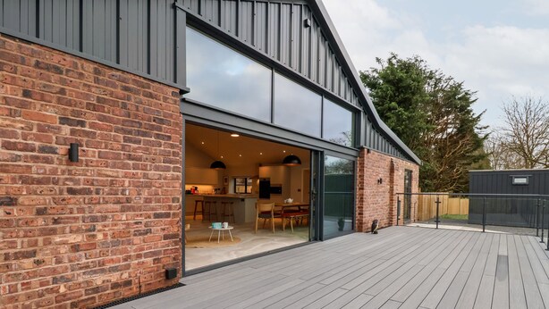 A modern building with a brick facade, large sliding glass doors leading to an interior with a kitchen and dining area, and a wooden deck. - Grange Barn (Shrewsbury)