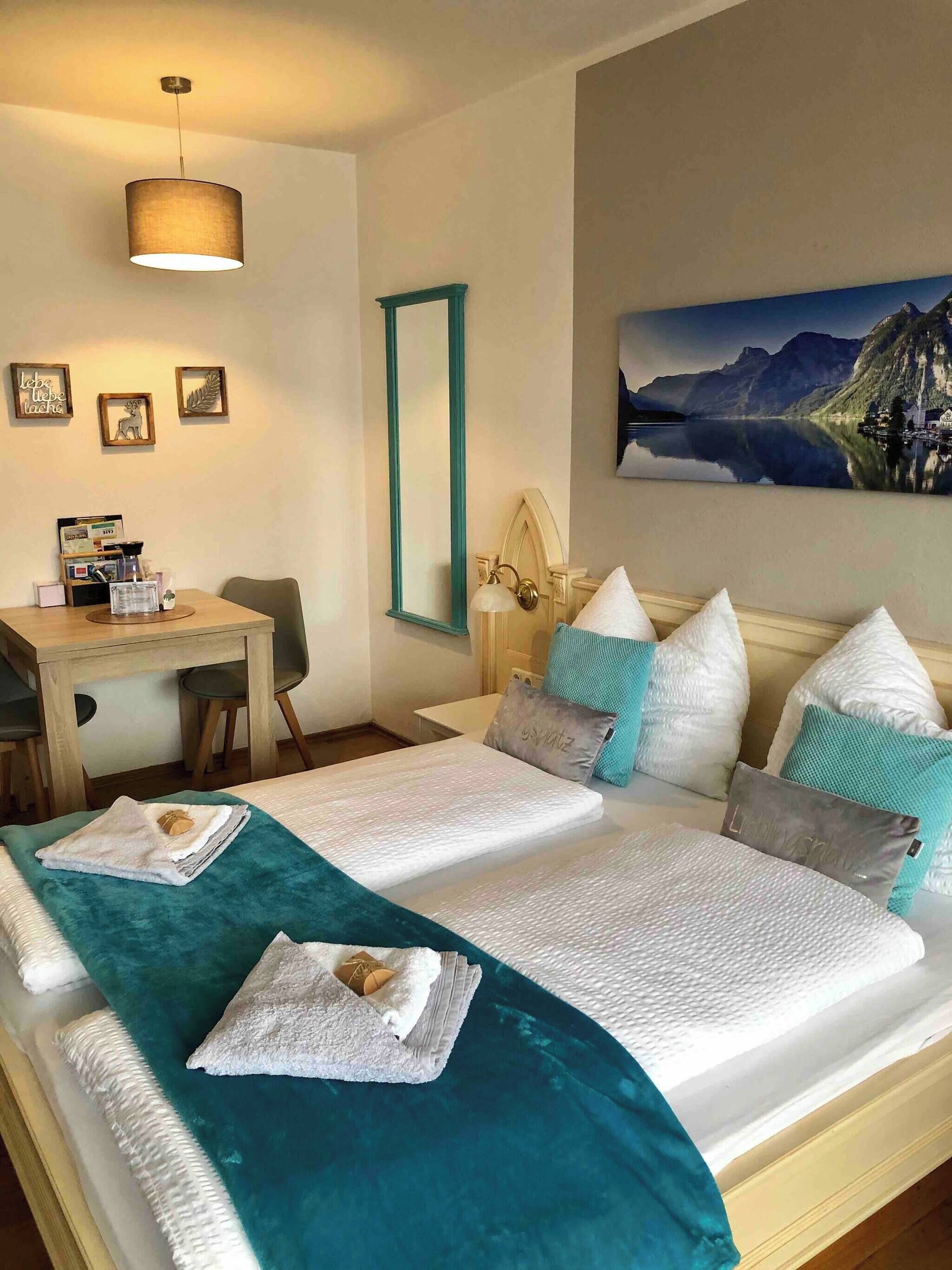 A hotel room with a bed, a desk, a chair, a lamp, and a painting of a mountain landscape.