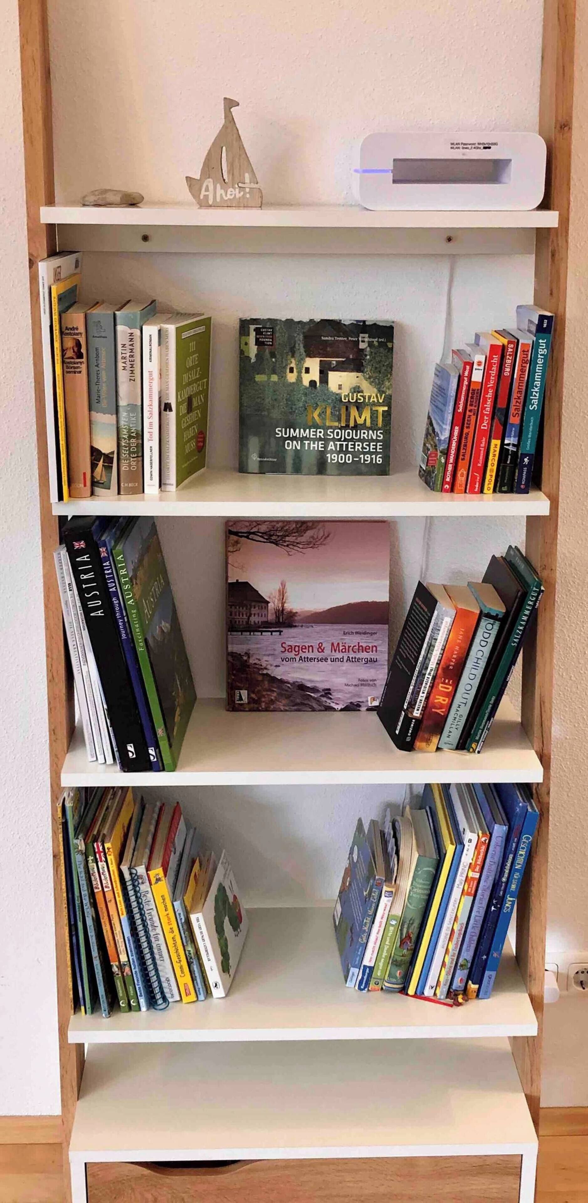 Une étagère remplie de divers livres, une petite décoration en forme de bateau en bois et un appareil électronique blanc.