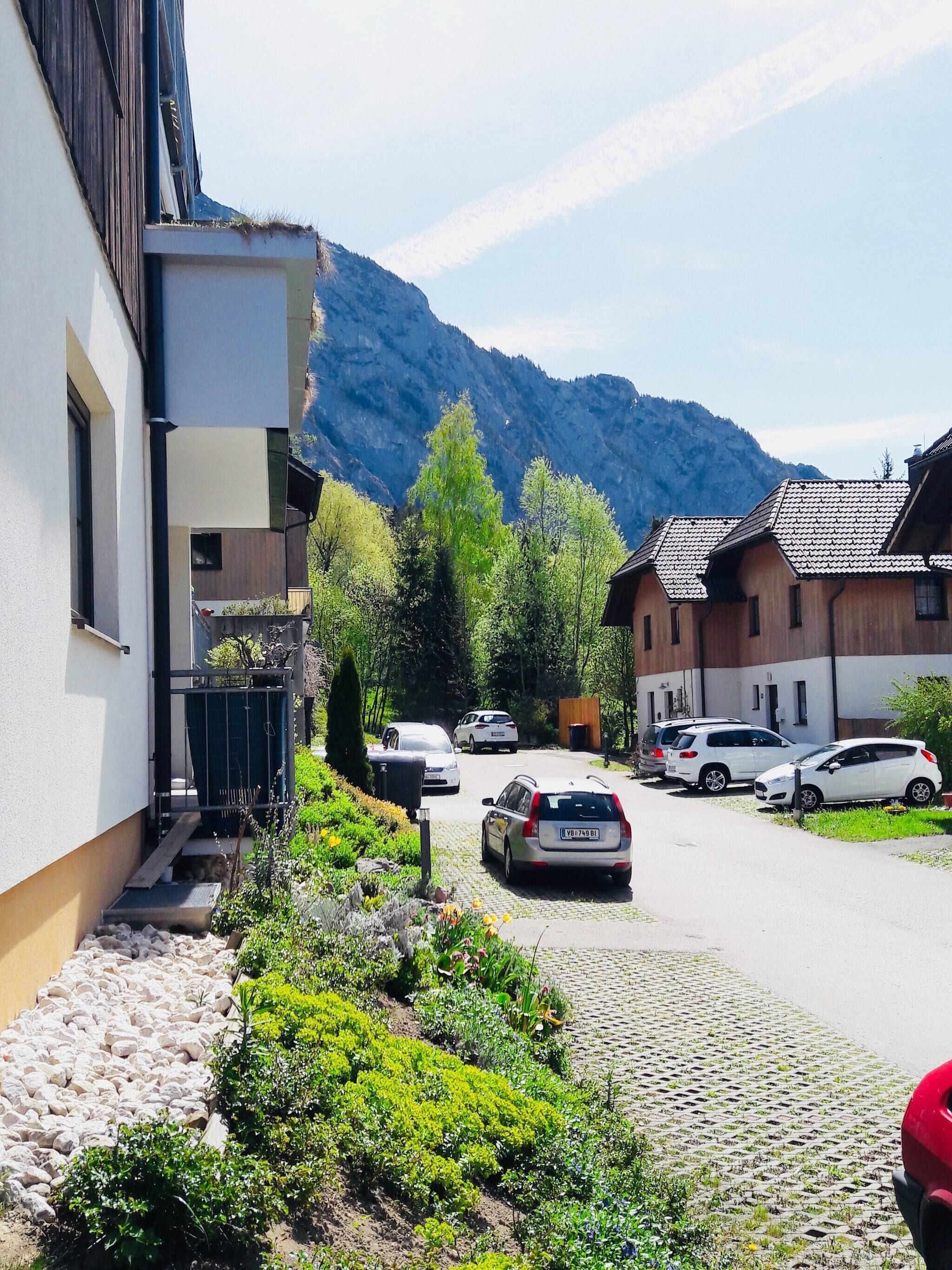 A residential street with parked cars, a building with a balcony, and a mountain in the background.