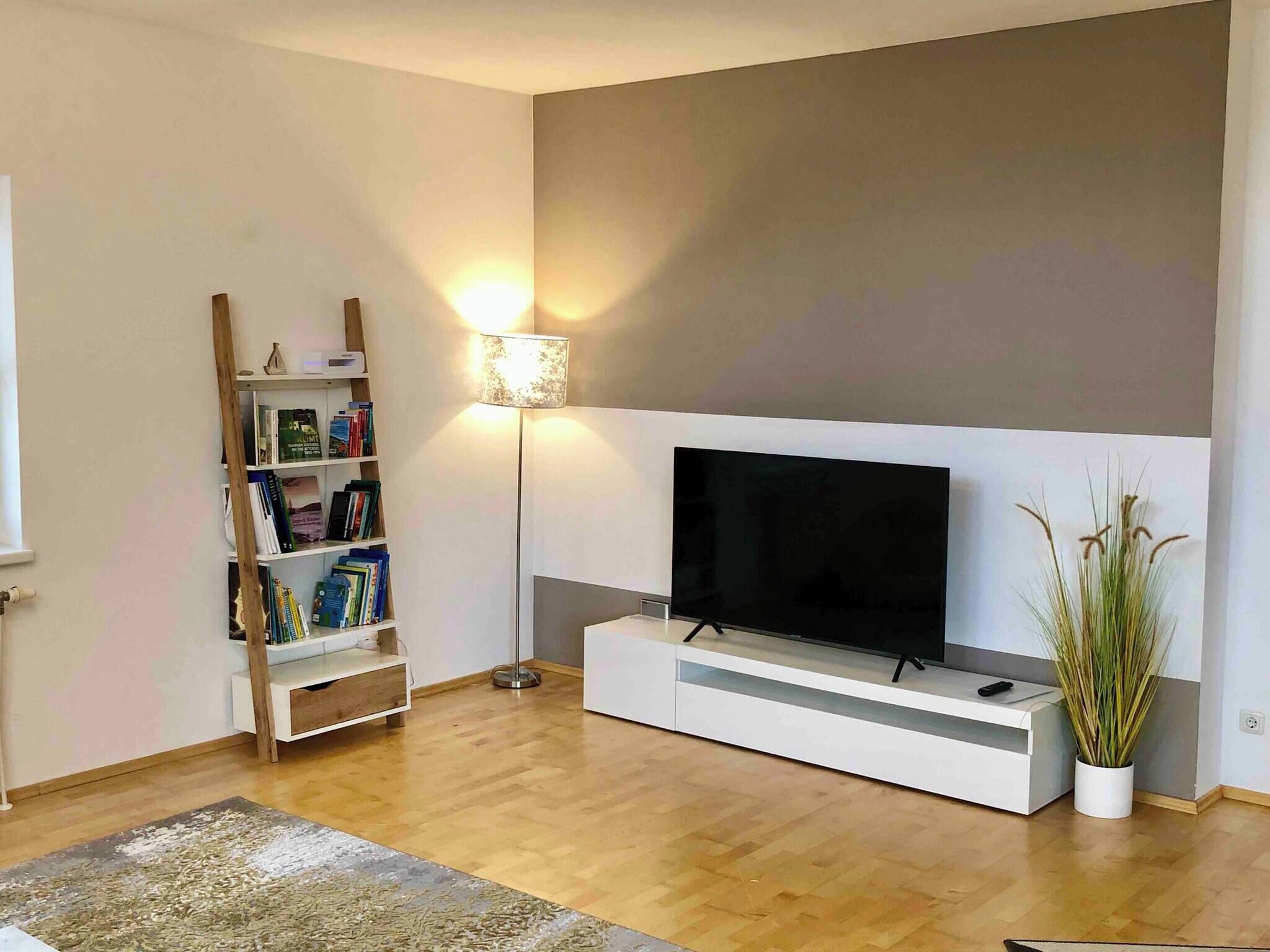 A living room with a flat-screen TV on a white stand, a bookshelf, a floor lamp, and a potted plant.