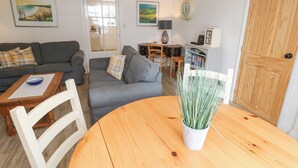 A cozy living room with a grey sofa, a wooden dining table, and a white chair. There is a small table with a bowl on it, a lamp, and a bookshelf. - Kelsey (Truro)