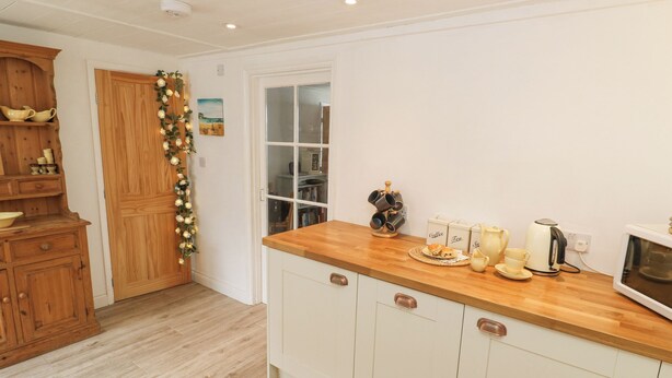 A kitchen with wooden cabinets, a wooden countertop, and a microwave. There is a coffee maker, a kettle, and a teapot on the counter. - Kelsey (Truro)