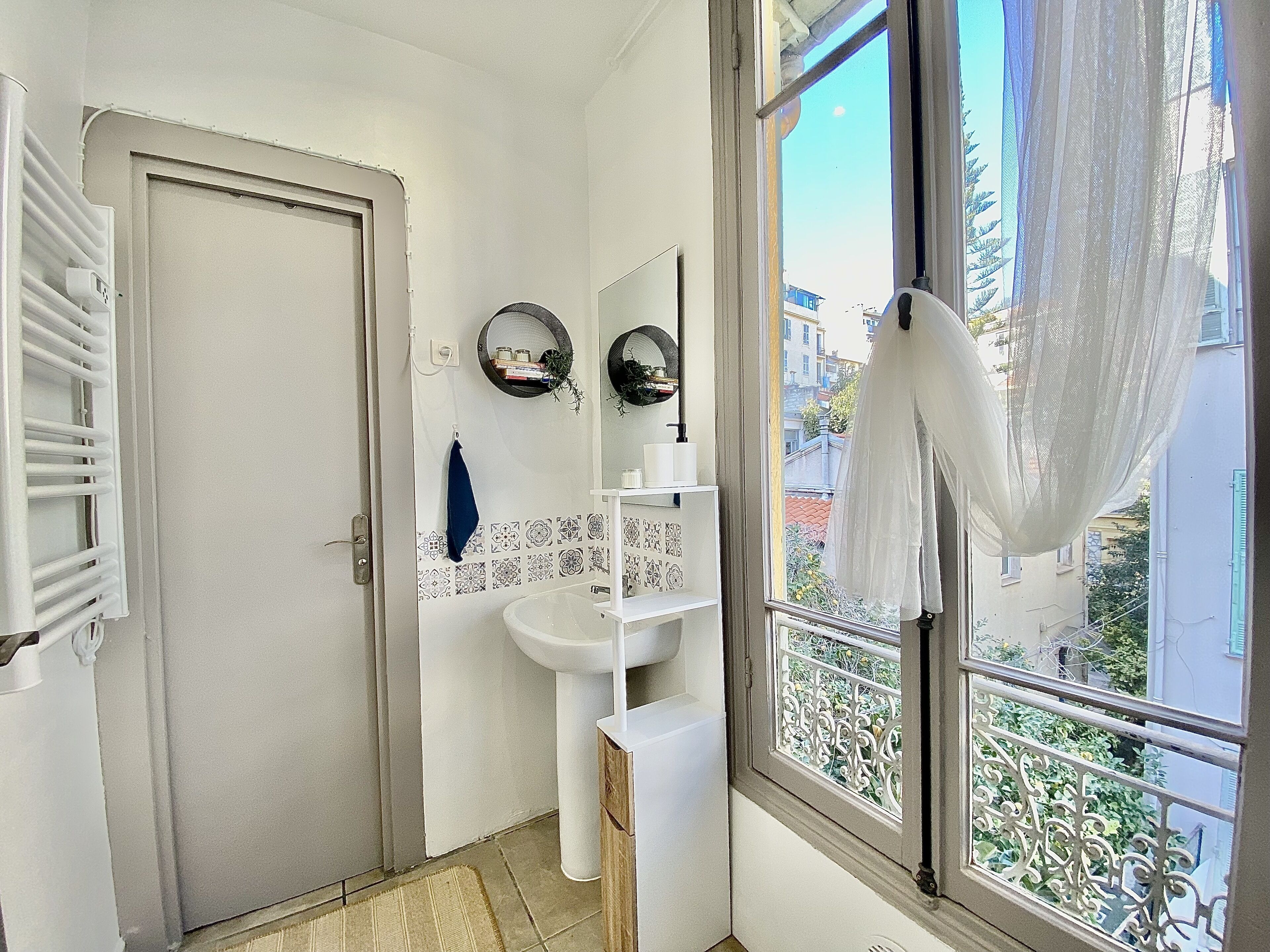 A bathroom with a white door, a sink, a mirror, and a window with a view of buildings and greenery.