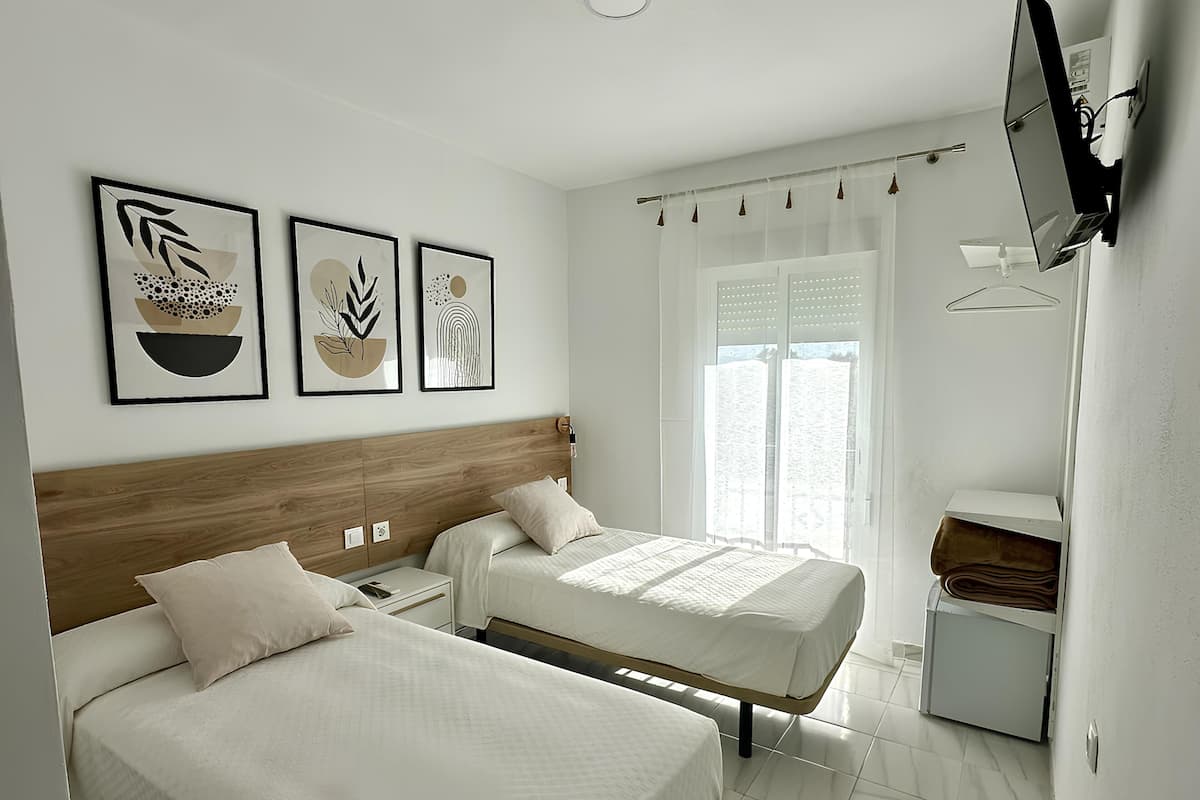 A hotel room with two beds, a wooden headboard, a TV mounted on the wall, and a window with sheer curtains.