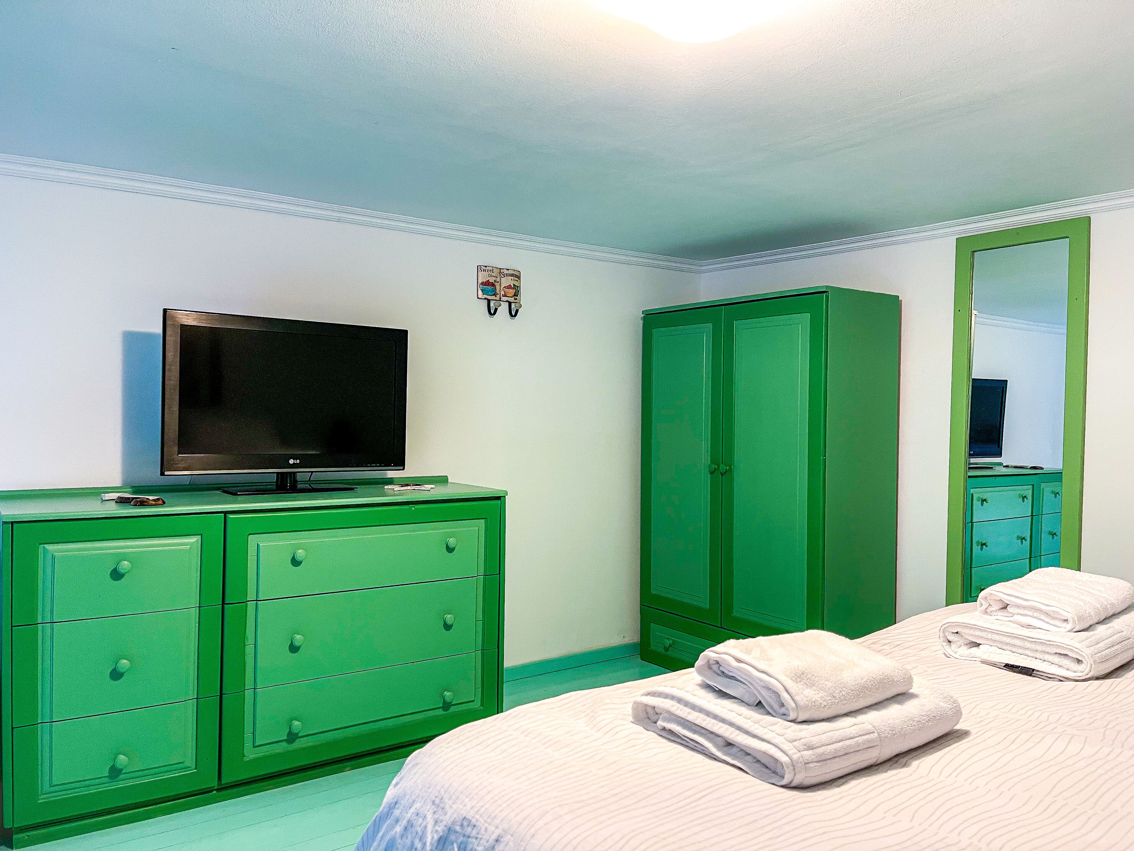 Un dormitorio con un aparador verde, un televisor de pantalla plana, un armario y una cama con toallas dobladas.