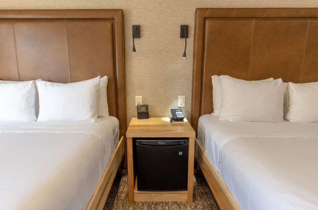 A hotel room with two beds, a small wooden side table with a phone and a remote control, and a compact refrigerator.