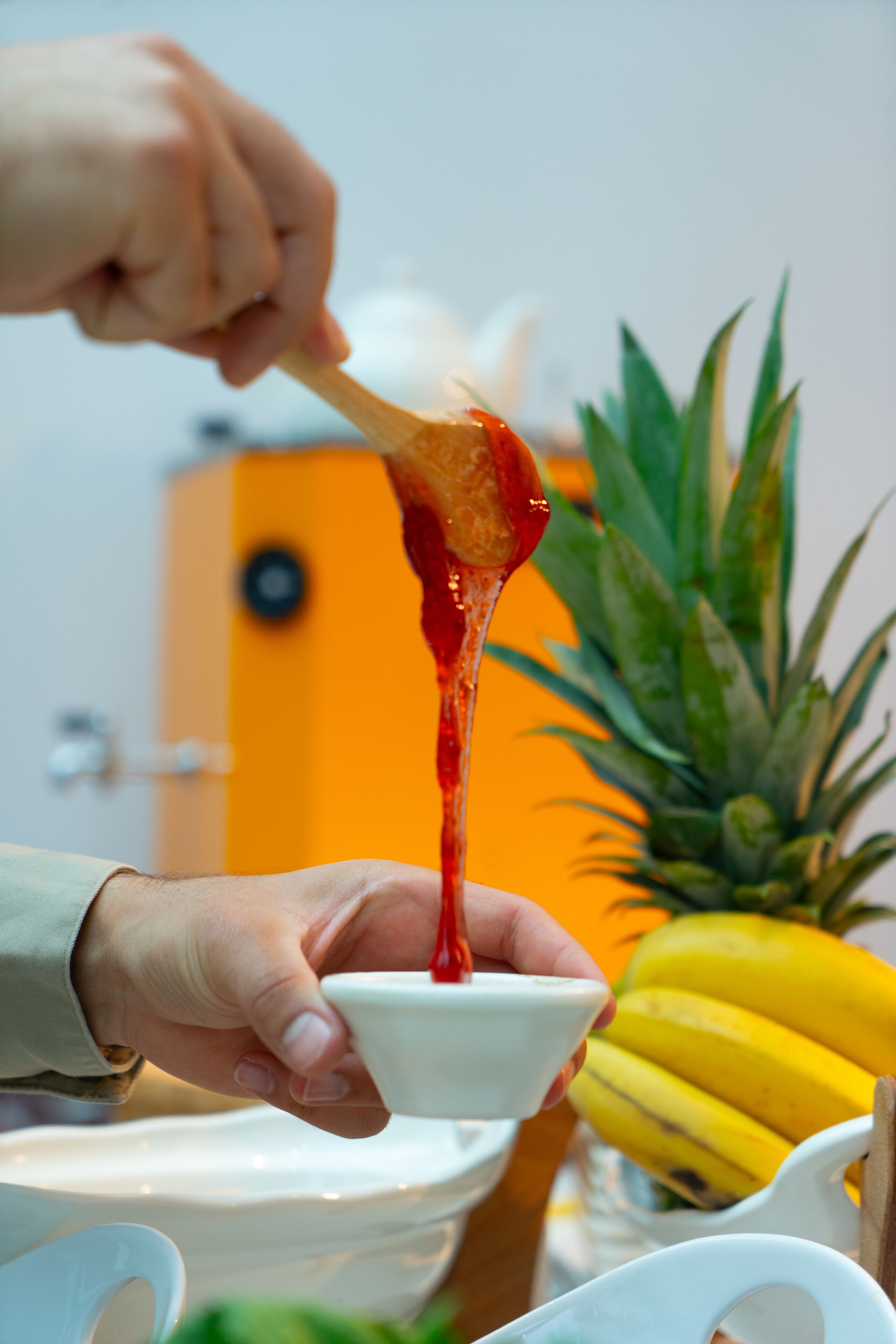 Une personne tient une cuillère remplie d’une sauce rouge au-dessus d’un bol blanc. On aperçoit des bananes et un ananas à l’arrière-plan.