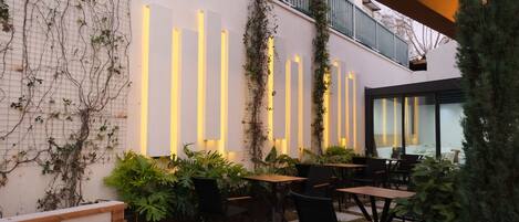 Un área de estar al aire libre con mesas y sillas, un banco de madera y una pared con paneles de luz verticales.