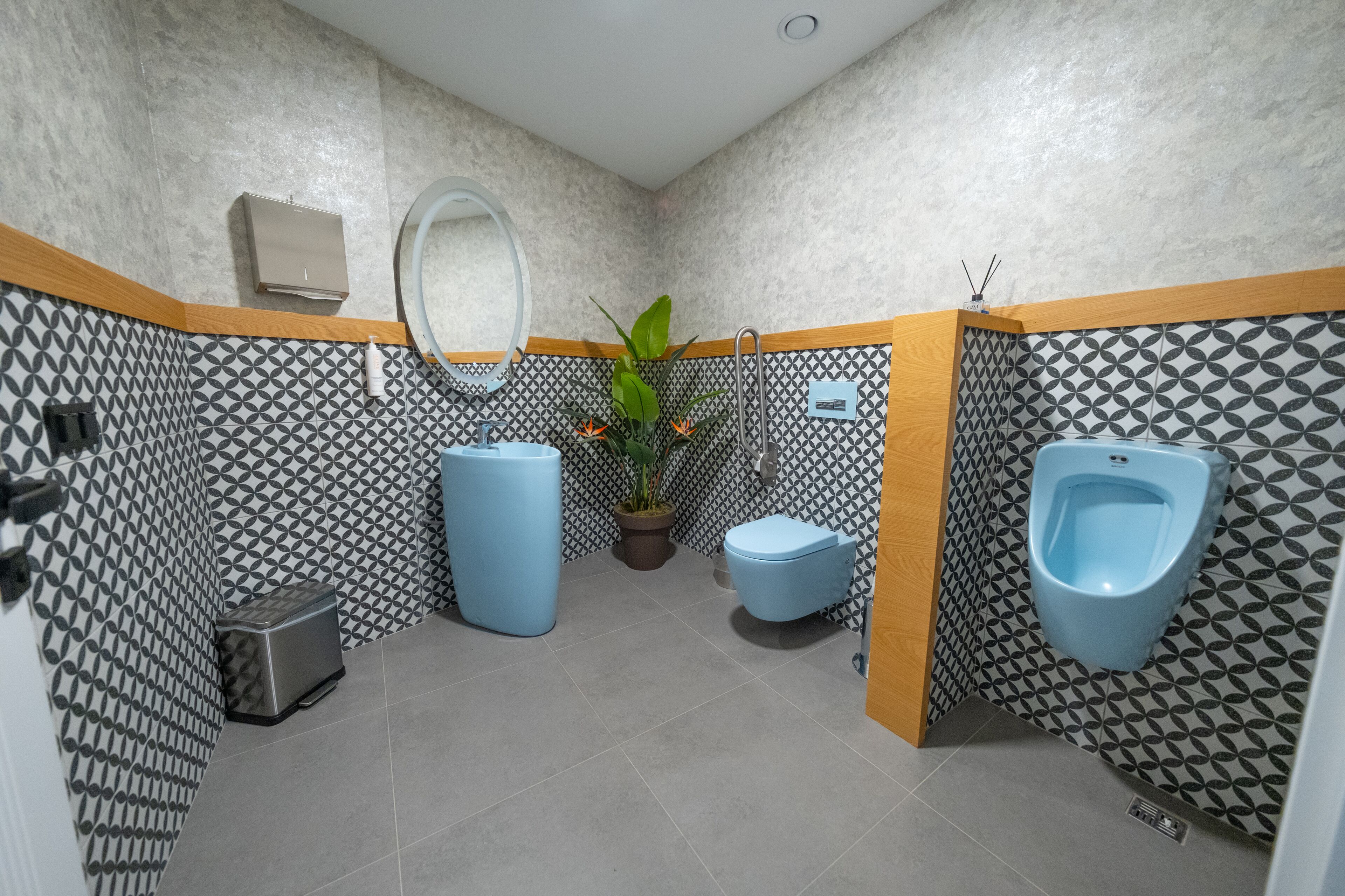 Een badkamer met een wand met een geometrisch patroon, een wastafel, toilet en urinoir, en een afvalbak.