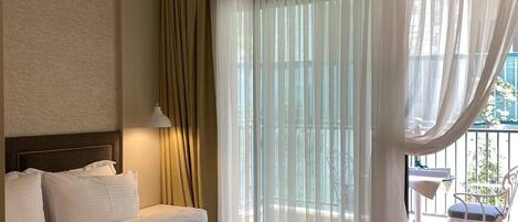 A hotel room with a large bed, a headboard, a bedside lamp, a window with sheer curtains, and a view of the outside balcony.