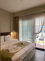 Habitación de hotel con una cama grande, cabecero, lámpara de noche, ventana con cortinas traslúcidas y vista al balcón exterior.