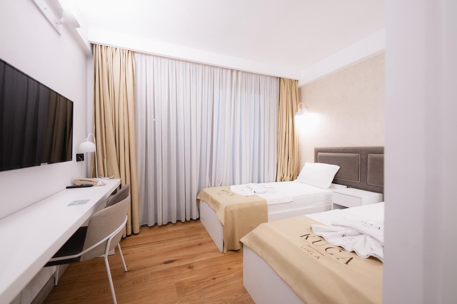 A hotel room with a desk, two beds, a TV, and a window with curtains.