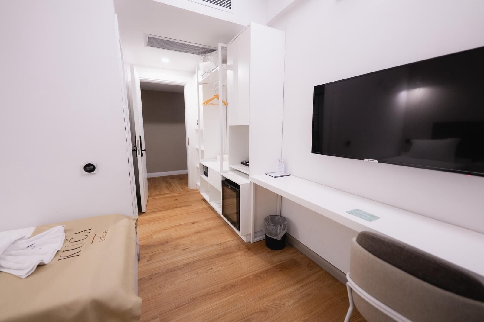 A modern hotel room with a wooden floor, white walls, a TV mounted on the wall, a wardrobe, and a small trash bin.