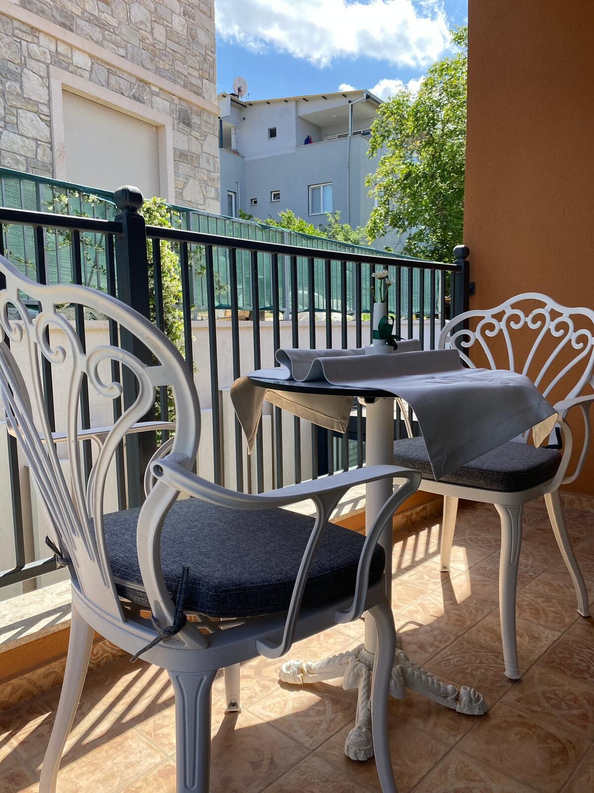 A balcony with two white metal chairs and a small table, a tiled floor, and a view of a street and buildings.
