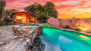 Une piscine dotée d’une plage en pierre et d’un espace jacuzzi, le tout entouré d’un jardin aménagé et surplombant un ciel au coucher du soleil.