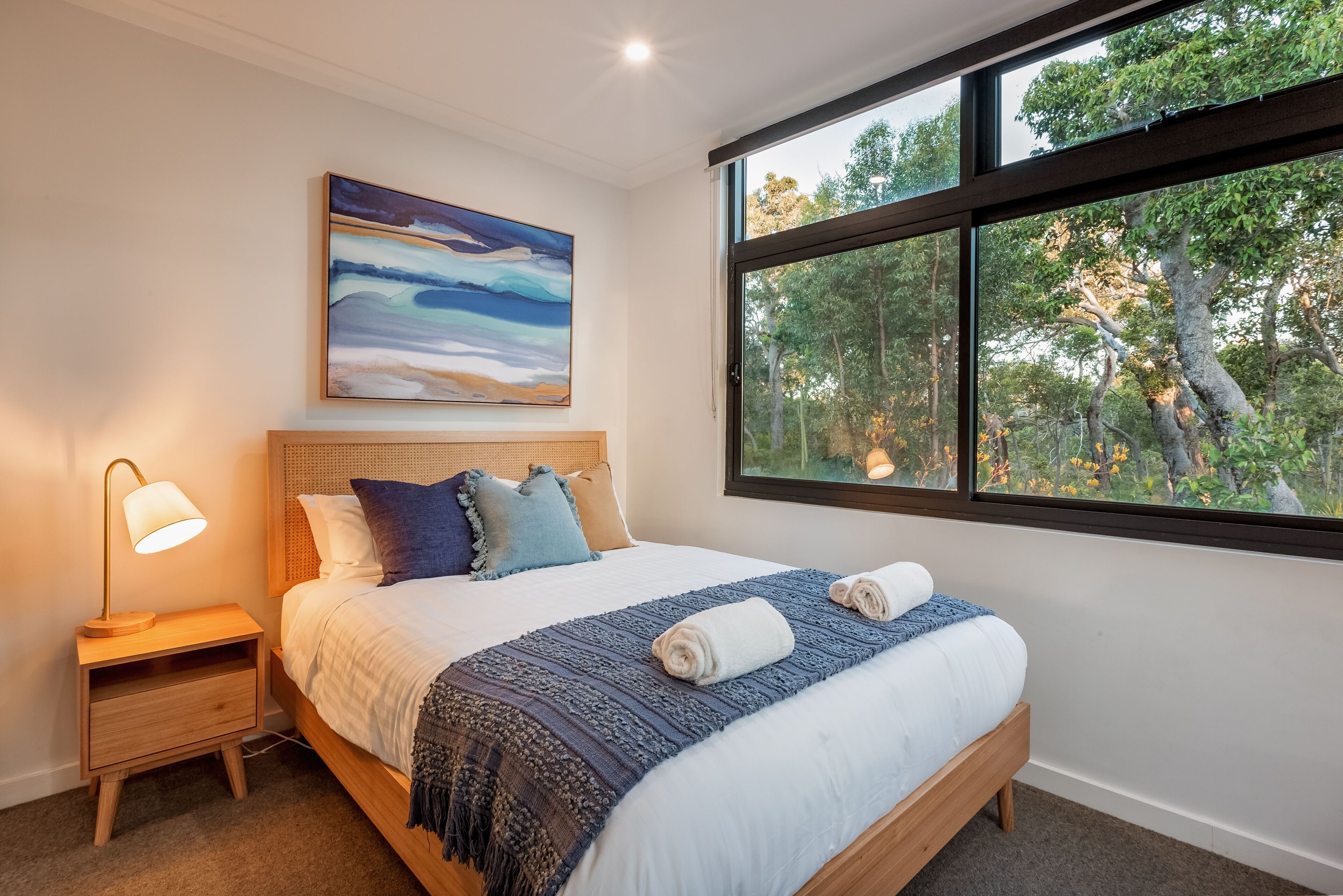 A bedroom with a large window, a bed with a blue blanket, a nightstand with a lamp, and a painting on the wall.
