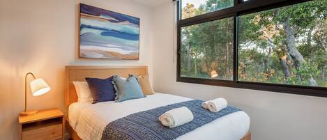 A bedroom with a large window, a bed with a blue blanket, a nightstand with a lamp, and a painting on the wall.