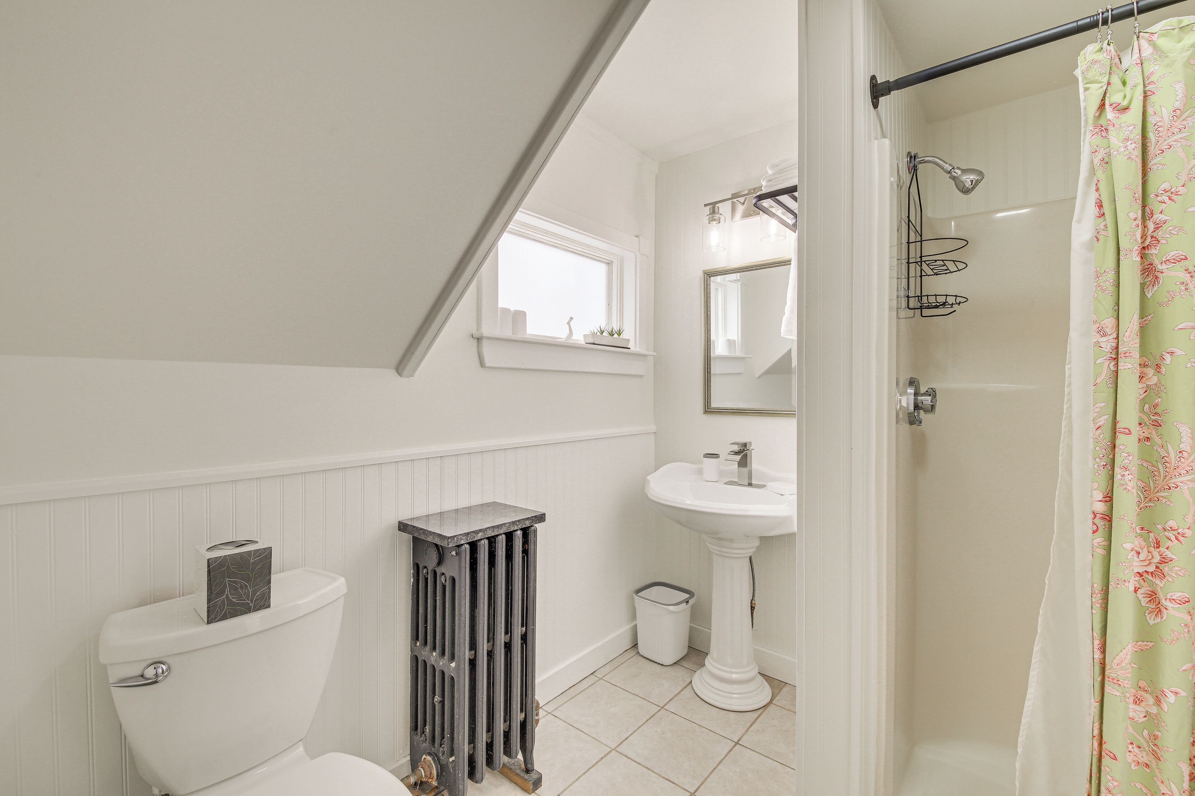 A bathroom with a toilet, pedestal sink, shower, and a window above the sink.