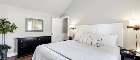 A bedroom with a large bed, a dresser, a ceiling fan, a mirror, and a nightstand with a lamp.