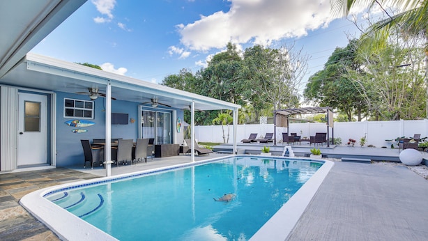 Ein Pool mit überdachter Terrasse, Gartenmöbeln und Blick auf Bäume und einen weißen Zaun.