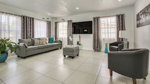 Ein modernes Wohnzimmer mit einem grauen Sofa, passendem Hocker und einem dunkelgrauen Sessel. Ein großes Fenster mit gemusterten Vorhängen, ein an der Wand befestigtes Flachbildfernsehen und eine Zimmerpflanze in der Ecke.