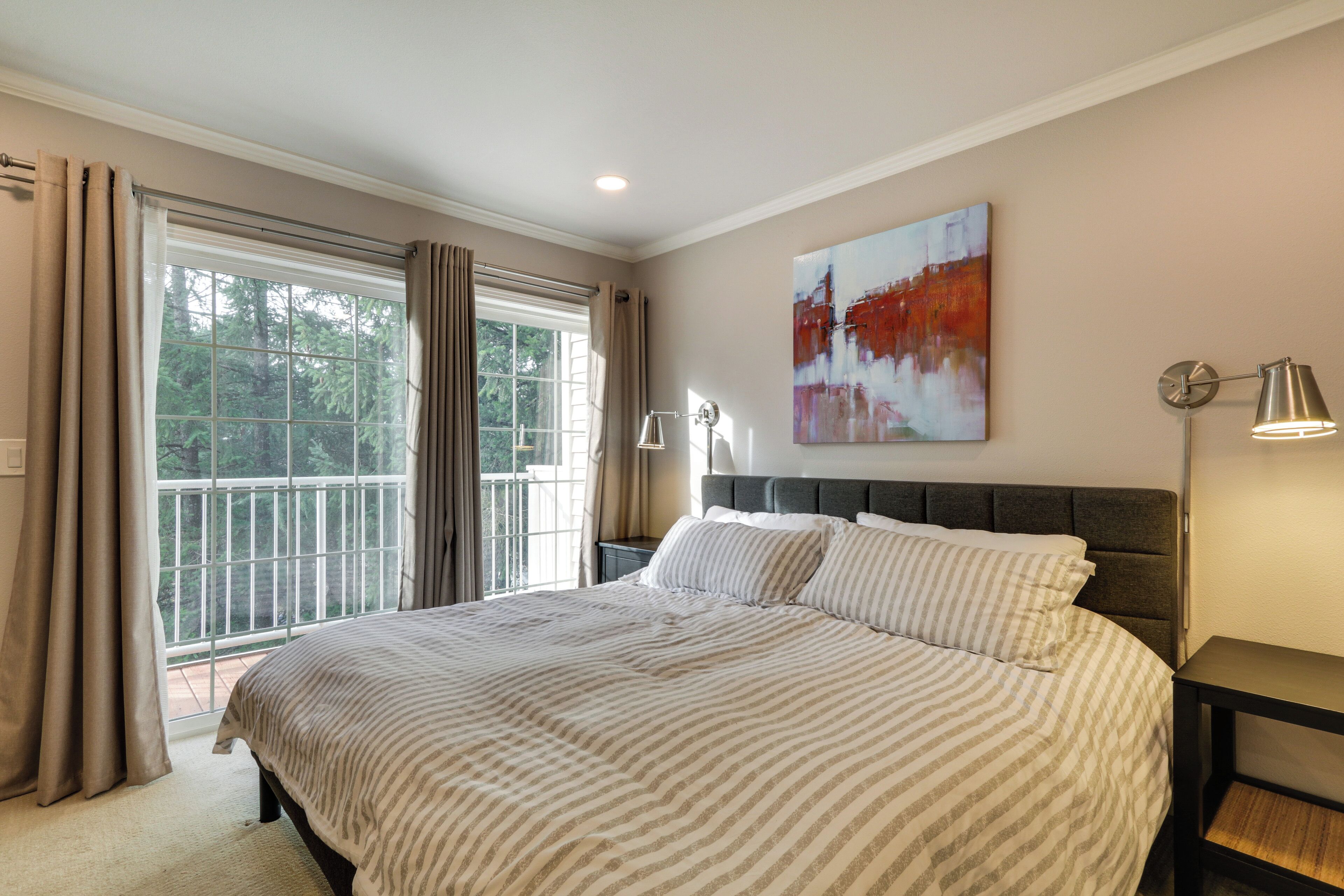 A bedroom with a large bed, a nightstand, a lamp, a painting on the wall, and a view of trees through the window.