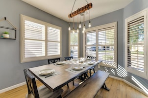 A dining area with a long table, chairs, and a bench. There are large windows with blinds, a hanging light fixture, and a small shelf with a plant.