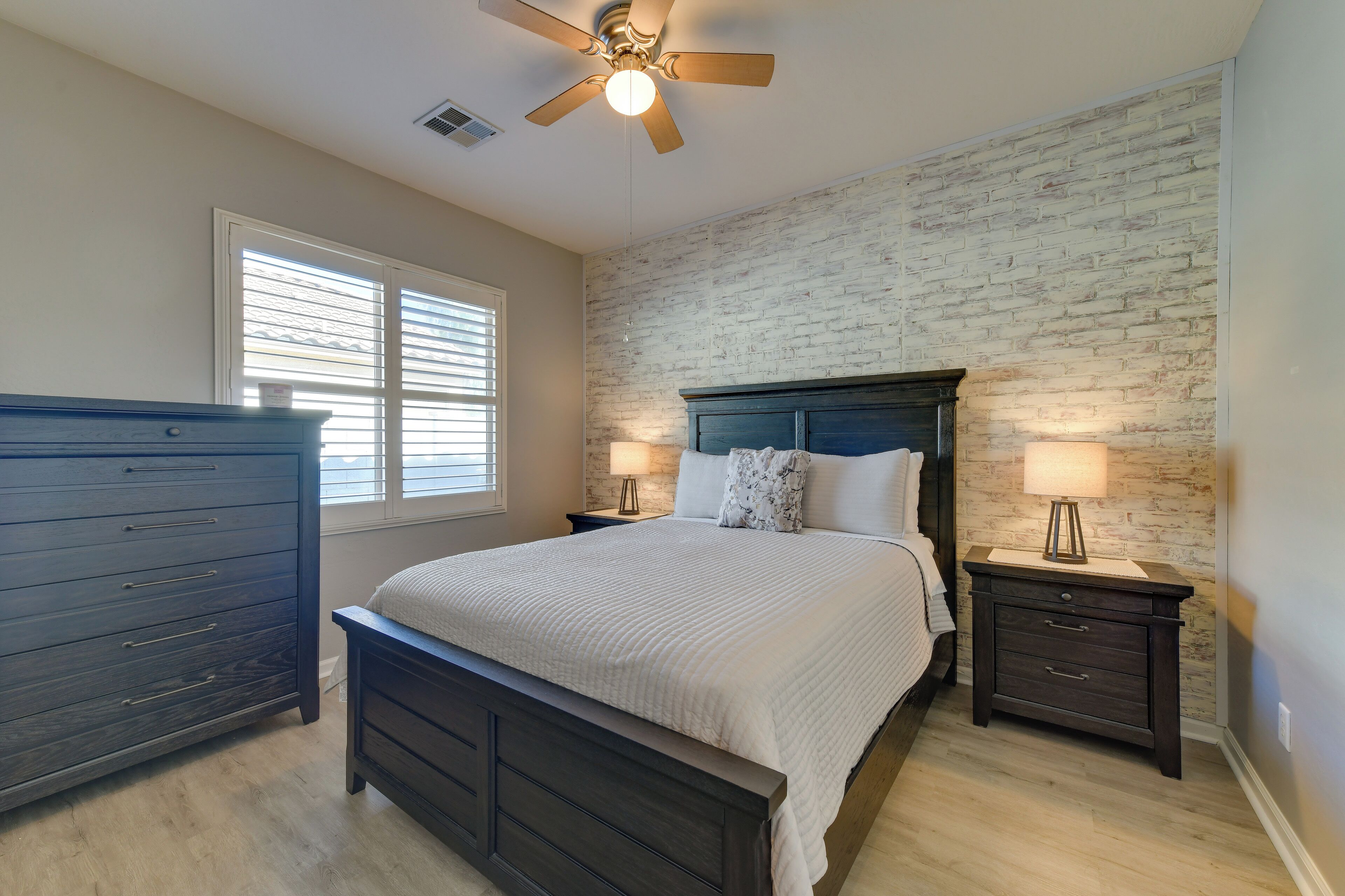 A bedroom with a dark wooden bed, a nightstand with a lamp, a dresser, and a brick-patterned wall.