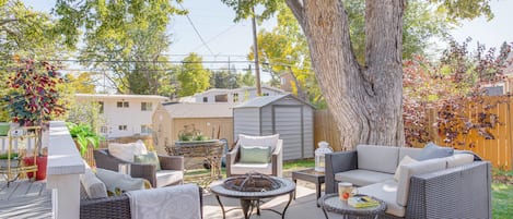 Un patio al aire libre con mobiliario de mimbre, una chimenea y un árbol grande que proporciona sombra.