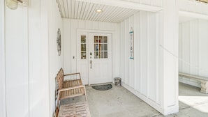 Une façade blanche, un banc en bois, une porte avec une fenêtre et une pancarte indiquant « Bienvenue ».